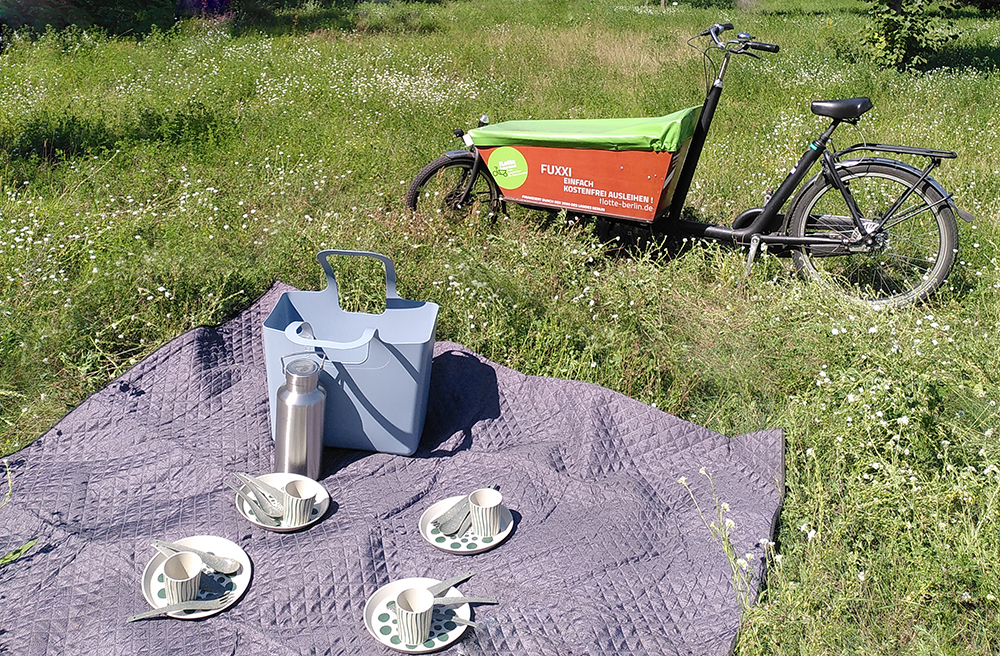 Wiese mit Picknickdecke, Teller und Tassen, im Hintergrund ein Lastenrad